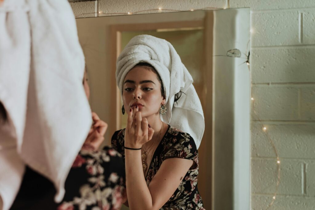 Woman with towel around her head applying makeup in a bathroom mirror